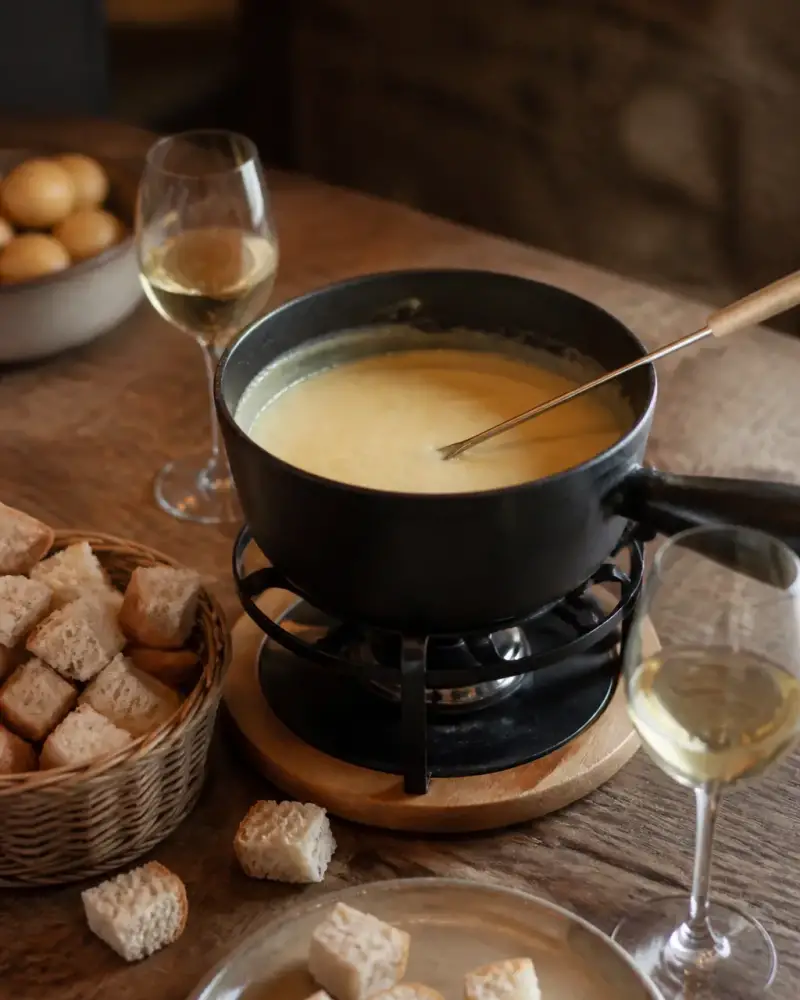 Caquelon de fondue au fromage sur un réchaud, cubes de pain et verres de vin blanc sur une table en bois