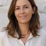 Portrait de Claire Montferrand, cheveux châtains mi-longs, chemise en lin claire, léger sourire, mur de pierre claire flou en arrière-plan