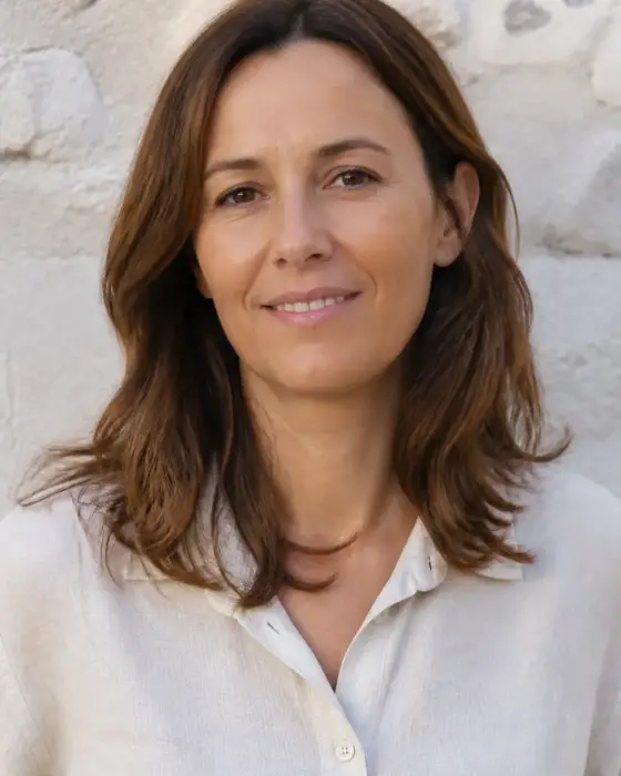 Portrait de Claire Montferrand, cheveux châtains mi-longs, chemise en lin claire, léger sourire, mur de pierre claire flou en arrière-plan