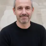 Portrait d’Étienne Roubaud, cheveux gris courts, barbe courte, pull sombre, sourire discret, mur de pierre claire flou en arrière-plan