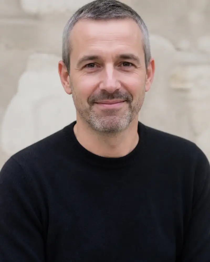 Portrait d’Étienne Roubaud, cheveux gris courts, barbe courte, pull sombre, sourire discret, mur de pierre claire flou en arrière-plan