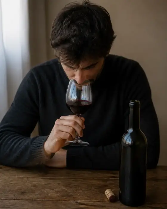 Verre de vin rouge sous le nez d’une personne qui hume, bouteille ouverte et bouchon posés sur une table